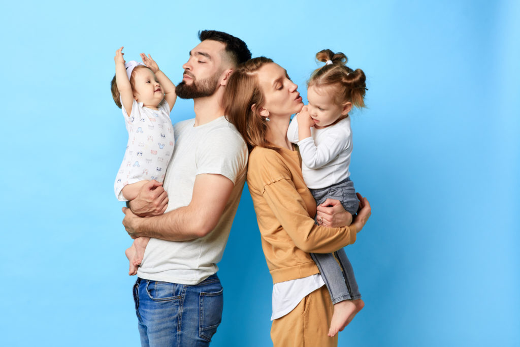 two parents standing back to back and holding their children in arms. close up photo. isolated blue background. studio shot.